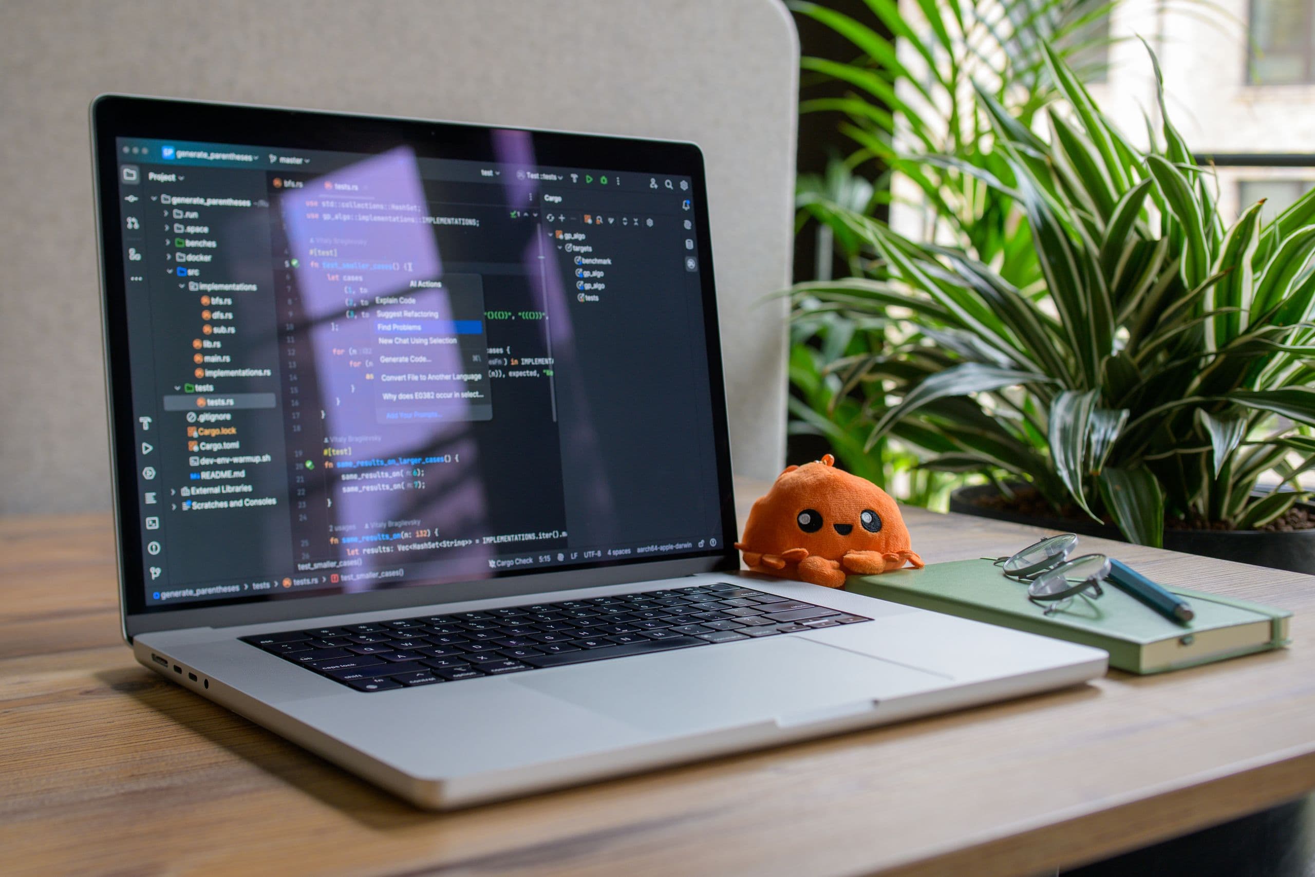 An open laptop on a wooden desk showing a code editor, with a green plant, a notebook and a small plush toy beside it in daylight
