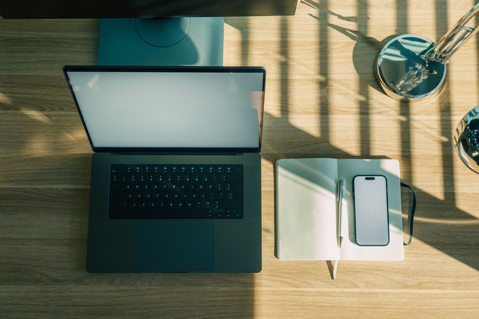 Top-down view of a sunlit wooden desk with an open laptop, a notebook, a pen and a phone